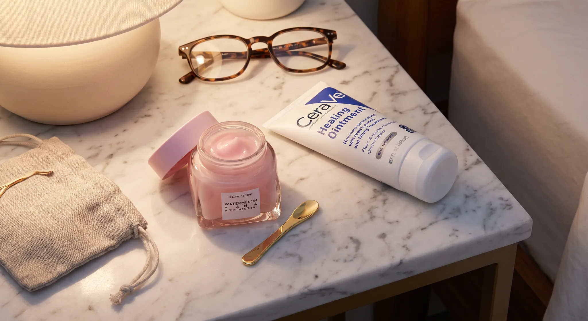 A detailed flat lay photography shot of nighttime skincare essentials: a jar of overnight hydrating mask, a spatula, and a tube of thick barrier ointment for slugging.