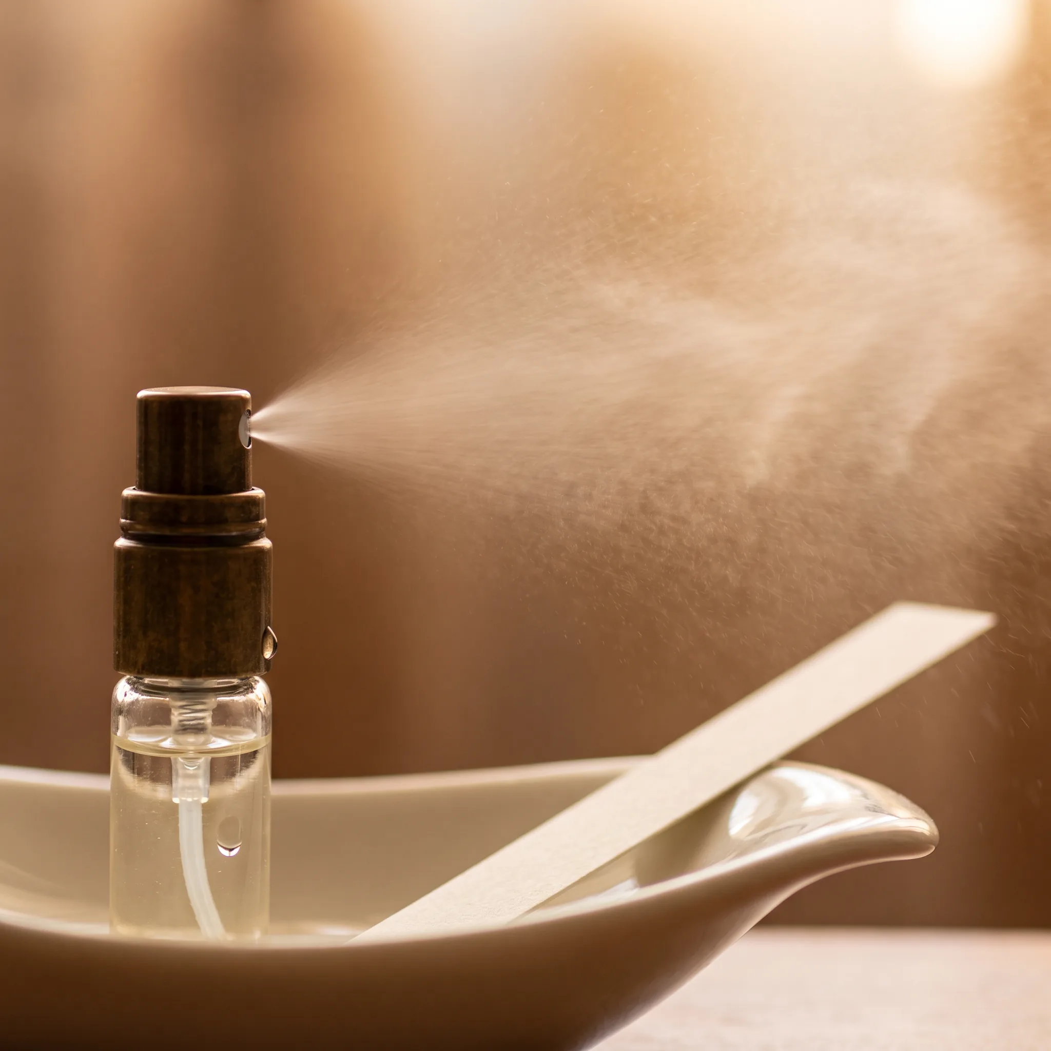 Close macro still life of a small glass travel spray releasing a fine fragrance mist into still air above a warm porcelain dish. A single pale paper b