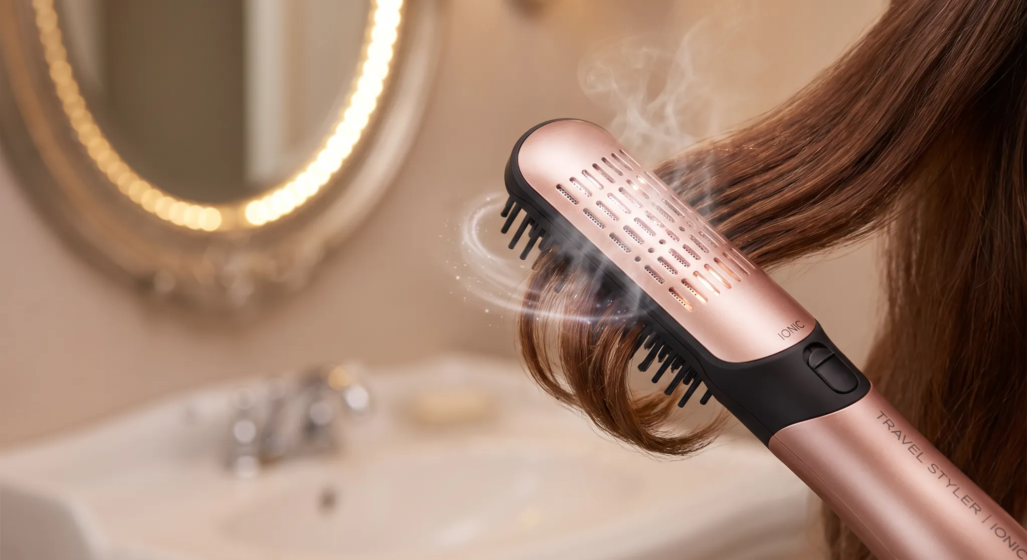 Close-up beauty demonstration photography showing a compact travel hair dryer brush smoothing and curling under the ends of lustrous hair in front of a warm hotel vanity mirror.