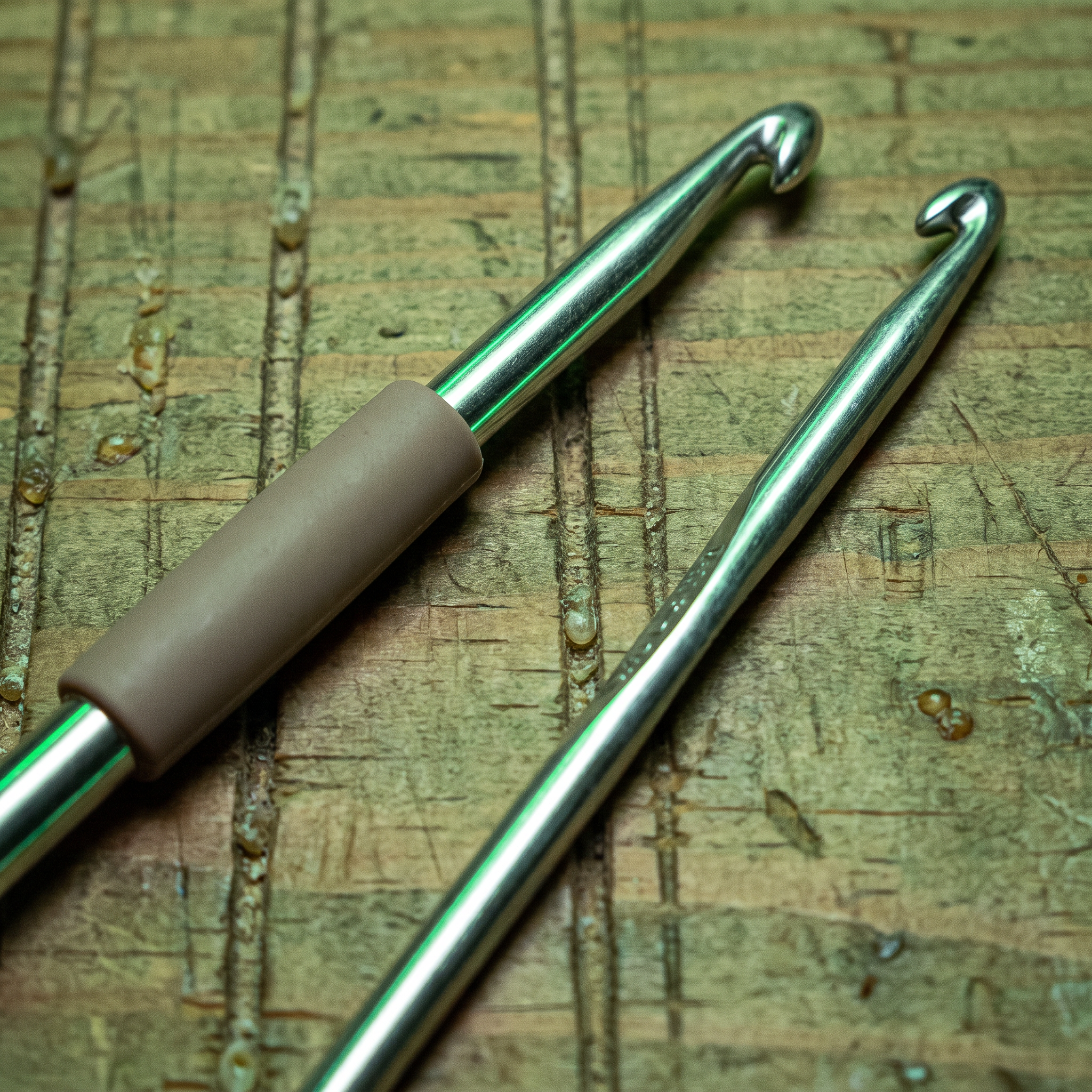 Two aluminum crochet hooks on a scored pine table, one bare and one with a taupe rubber grip sleeve halfway on